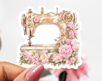 May include: A sticker featuring a vintage sewing machine design. The machine is cream-colored with gold accents and adorned with pink floral arrangements. The sticker is held by a hand against a soft background.