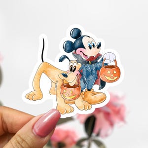 May include: A sticker featuring Mickey Mouse dressed as a vampire and Pluto holding jack-o'-lantern treat buckets. Mickey wears a blue suit and cape. Pluto is light brown. The sticker has a white border and is held by a hand.