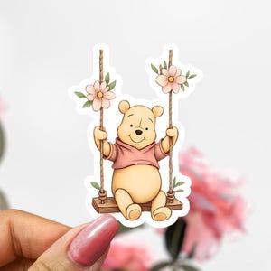 May include: A sticker depicting a cartoon bear in a pink hoodie on a wooden swing. The swing is decorated with pink flowers and green leaves. The bear is smiling, and the design has a whimsical, cheerful aesthetic. The image is a sticker.