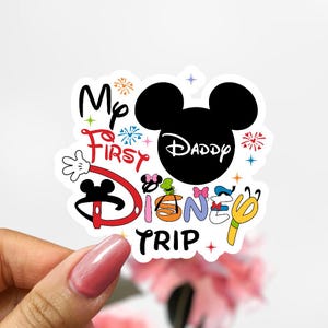 May include: A white sticker featuring the phrase "My First Disney Trip" in a playful, colourful design. The design incorporates a black Mickey Mouse head silhouette with the word "Daddy" inside, along with decorative elements like fireworks and character details.