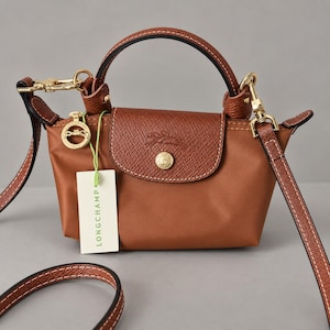May include: A small brown handbag with a top handle and a detachable shoulder strap. The bag features a textured leather flap with a gold-tone snap button, a gold-tone logo medallion, and a white tag with the text LONGCHAMP attached by a green string.