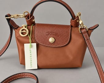 May include: A small brown handbag with a top handle and a detachable shoulder strap. The bag features a textured leather flap with a gold-tone snap button, a gold-tone logo medallion, and a white tag with the text LONGCHAMP attached by a green string.