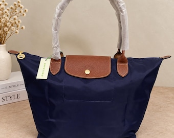 Peut inclure: Un sac cabas bleu marine doté d'un rabat en cuir marron avec un bouton-pression doré. Le sac possède deux anses enveloppées de papier de protection blanc et une étiquette Longchamp attachée sur le côté. Le modèle inclut une fermeture éclair.