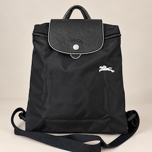 Op de afbeelding: Een zwarte nylon rugzak met een gestructureerde leren klep en een zilverkleurige drukknoopsluiting. De klep heeft een in reliëf gemaakt paardenlogo. Een wit geborduurd paardenlogo is zichtbaar op de voorzijde. Inclusief handvat en schouderbanden.