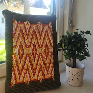 May include: A rectangular olive green crochet pouch featuring a central panel with a geometric zigzag pattern in vibrant shades of orange, yellow, and white. The pouch has a zippered top closure and a textured, dense stitch construction throughout.