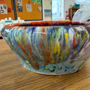 May include: A wide, round ceramic bowl featuring a vibrant, multi-colored glaze with swirling patterns of blue, yellow, orange, and white. The glossy finish creates a fluid, marbled effect across the exterior surface, tapering toward a flat base.