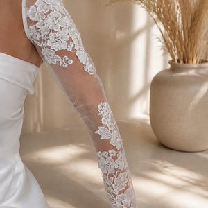 Peut inclure: Une manche de mariée longue et blanche en tissu maille transparent ornée de broderies florales blanches complexes. La manche s'étend de l'épaule au poignet, présentant un motif botanique détaillé qui recouvre le bras dans un style ajusté.
