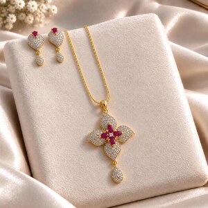 May include: A gold-tone jewellery set consisting of a necklace and two matching earrings. The necklace features a flower-shaped pendant with four marquise-cut red stones and clear pavé crystals, plus a small teardrop-shaped drop pendant.