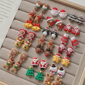 Pode incluir: Uma coleção de pequenos brincos de tarracha com temas natalinos, incluindo Papai Noel, renas, bonecos de gengibre, bonecos de neve, pinguins, estrelas e árvores de Natal, dispostos em uma bandeja de veludo bege para joias.
