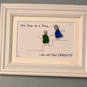 May include: A white framed picture with a hand-drawn illustration of two figures walking up a staircase. The figures are made of colorful glass stones. The text reads "One Step at a Time... we will find SERENITY".
