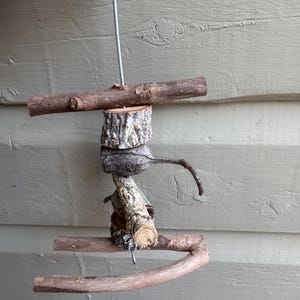 May include: A hanging bird toy constructed from natural wooden branches and bark-covered segments threaded onto a central metal rod. The structure features horizontal perches at the top and bottom, with various textured wood pieces in between.