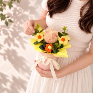 May include: A small pink pot containing a crocheted bouquet with a peach-colored bear head, two yellow flowers with orange centers, one orange flower with a yellow center, and green leaves. The pot is wrapped in yellow paper and tied with a cream ribbon.