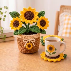 Puede incluir: Una planta de girasol de ganchillo en una maceta marrón con borde y lazo color crema, acompañada de una taza de cerámica blanca con estampado de girasol, sobre un posavasos de girasol de ganchillo a juego.
