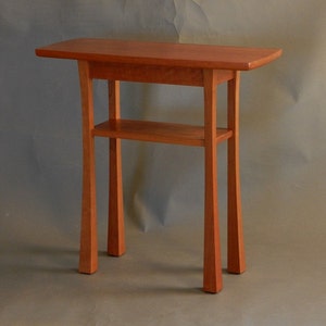 May include: A small, wooden table with a single shelf. The table is made of cherry wood and has a natural finish. The table is about 30 inches tall and 24 inches wide.