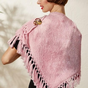 May include: A triangular knit shawl in a soft pink hue, featuring a textured stitch pattern and long fringe along the two bottom edges. The shawl is secured at the shoulder with a decorative gold-toned brooch featuring colorful enamel accents.