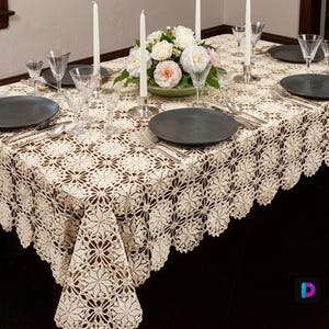 Peut inclure: Une nappe rectangulaire en crochet de couleur crème présentant un motif floral répétitif avec des bords festonnés. La nappe est drapée sur une table dressée avec des assiettes noires, de la verrerie transparente, des couverts en argent, des bougies blanches et un centre de table floral.