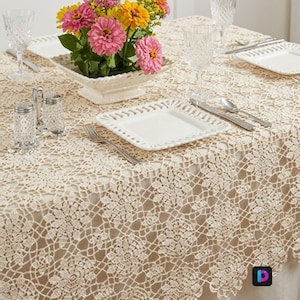 May include: A cream-coloured crochet tablecloth with a floral pattern draped over a table. The table is set with white square plates, clear crystal glasses, silver cutlery, salt and pepper shakers, and a centrepiece of pink and yellow flowers.
