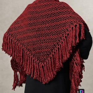 May include: A deep red triangular shawl featuring an openwork crochet pattern and long, thick fringe along the bottom edges. The garment is draped over the shoulders, showcasing the intricate yarn texture and the consistent dark crimson color throughout.