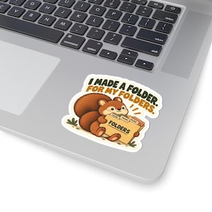 May include: A die-cut sticker featuring a brown squirrel holding a tan file folder labelled FOLDERS, with smaller folders inside. The text above the squirrel reads I MADE A FOLDER. FOR MY FOLDERS. in dark brown, rounded, sans-serif capital letters.
