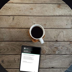 Puede incluir: Una taza de cerámica blanca llena de café negro sobre un posavasos de madera junto a una tableta que muestra un documento titulado Food Safety Management System. La pantalla muestra texto sobre procedimientos operativos estándar.