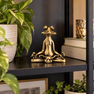 Puede incluir: Una figura de rana de color oro metálico en una pose de meditación sentada con las piernas cruzadas y las manos descansando sobre las rodillas. La rana tiene una superficie lisa y reflectante con detalles antiguos oscuros en las grietas.