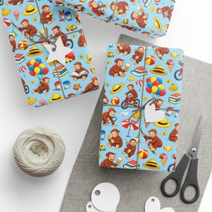 May include: Light blue wrapping paper featuring a repeating pattern of Curious George monkeys, yellow hats, red bicycles, colorful balloons, stacks of books, ice cream cones, and paint cans, tied with twine and white star-shaped gift tags.