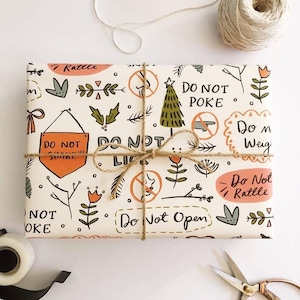 May include: A rectangular gift wrapped in cream-colored paper featuring a repeating pattern of botanical illustrations, pine trees, and orange warning signs with text including Do Not Poke, Do Not Shake, Do Not Lick, and Do Not Open, tied with brown twine.