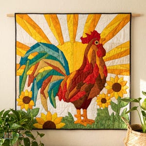 May include: A square fabric wall hanging featuring a quilted rooster in shades of brown and red, standing in a field of green with yellow sunflowers. The background displays a bright yellow sunburst pattern against a white sky.