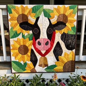 Puede incluir: Un tapiz acolchado que muestra una vaca blanca y negra con nariz rosa y una campana dorada, rodeada de grandes girasoles amarillos con hojas verdes. Las piezas de tela están unidas con líneas de acolchado visibles en todo el diseño.