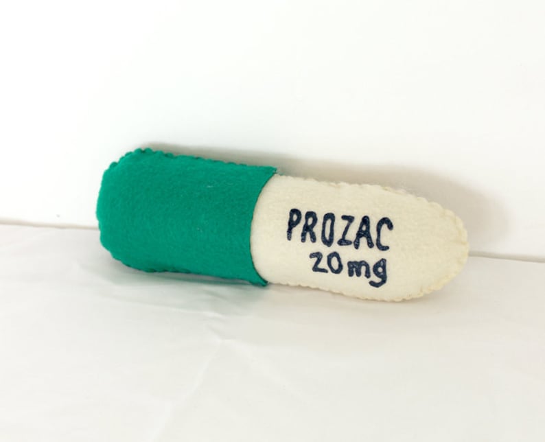 Puede incluir: Una pastilla de fieltro verde y blanco con forma de c&aacute;psula con el texto "PROZAC 20mg" bordado en el lado blanco.