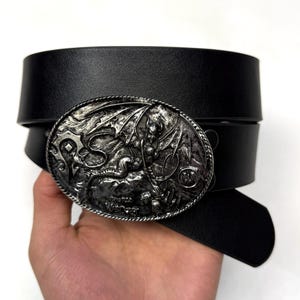 May include: A black leather belt featuring an oval-shaped silver-toned metal buckle. The buckle displays a detailed relief of a winged creature and the text World of Warcraft, with a rope-style border surrounding the central embossed design.