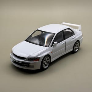 Może przedstawiać: Biały model samochodu typu sedan Mitsubishi Lancer Evolution IX. Pojazd posiada tylny spojler, srebrne wieloramienne felgi oraz czarną przednią tablicę rejestracyjną z napisem Lancer Evolution IX na przednim zderzaku.