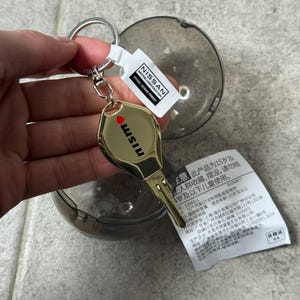 Peut inclure: Un porte-clés en métal doré en forme de clé de voiture, avec le texte noir 'nismo' et un point rouge. Il comprend un anneau fendu argenté et une étiquette blanche avec le texte 'NISSAN MOTOR CORPORATION OFFICIAL LICENSED PRODUCT'.