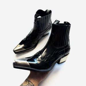 Puede incluir: Un par de botines negros con acabado brillante, que presentan una puntera metálica plateada y un tacón de bloque a juego. Los botines tienen paneles laterales elásticos y una trabilla negra en la parte superior con el texto J'ADIOR en blanco.