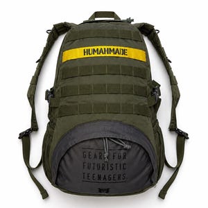 Puede incluir: Una mochila de color verde oliva con una correa horizontal amarilla que lleva el texto HUMANMADE en negro. El bolsillo delantero inferior es gris oscuro con cierre de cordón y el texto GEAR FOR FUTURISTIC TEENAGERS impreso en negro.