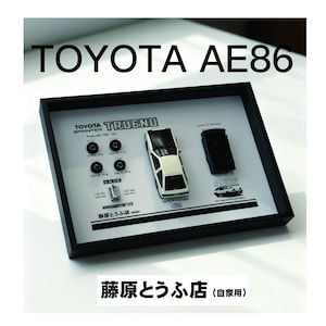 May include: A framed display featuring a white and black Toyota AE86 model car, four miniature wheels, and a small engine block. The white background includes technical text, diagrams, and the Japanese characters 藤原とうふ店. The frame is black.