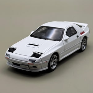 Può includere: Un modellino in scala bianco di un'auto sportiva Mazda RX-7 con fari a scomparsa, presa d'aria sul cofano, cerchi argentati a più razze e una targa con la scritta 13-954. L'auto presenta un logo rosso sul pannello posteriore e finiture nere.