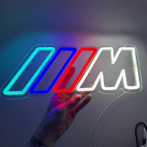 Pode incluir: Um letreiro de néon com o logótipo M, apresentando três barras inclinadas em azul claro, azul escuro e vermelho, seguidas por uma letra M branca. O letreiro está montado numa base de acrílico transparente e emite um brilho intenso.