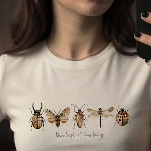 Może przedstawiać: Kremowy t-shirt z nadrukiem graficznym przedstawiającym pięć owadów, w tym chrząszcza, ćmę i ważkę, z wzorem tekstury starego papieru na skrzydłach i ciałach. Poniżej owadów znajduje się napis, the last of the bugs.