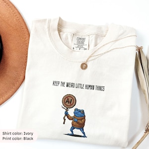 Pode incluir: Uma t-shirt cor de marfim com um gráfico preto de um sapo azul vestindo um suéter castanho e segurando uma placa de madeira com um círculo vermelho e uma barra sobre as letras AI. O texto acima do sapo diz KEEP THE WEIRD LITTLE HUMAN THINGS.