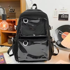 May include: A black backpack featuring two front pockets with clear plastic windows and silver D-ring hardware. The bag includes side compression straps with white buckles, a top handle, and a mesh panel visible behind the upper clear pocket.