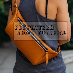 May include: A tan crossbody bag with a diagonal black zipper closure and a matching leather pull tab. The bag features the text PDF PATTERN VIDEO TUTORIAL printed across the center in a bold, dark font.