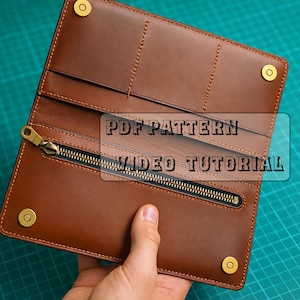 Peut inclure: Un portefeuille long en cuir marron doté d'un compartiment zippé, de plusieurs fentes pour cartes et de quatre boutons-pression en métal doré. Le portefeuille présente des coutures visibles et le texte PDF PATTERN VIDEO TUTORIAL.