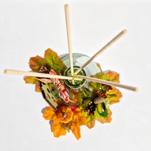 May include: A glass reed diffuser bottle with four light-colored wooden reeds inserted into the opening. The base is decorated with artificial autumn foliage, including orange and yellow maple leaves, small pinecones, an acorn, and a pumpkin.