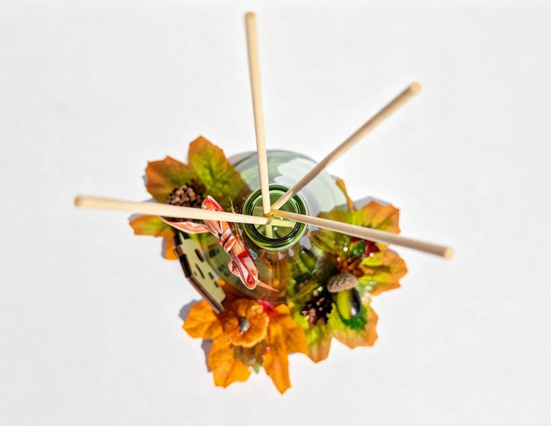 May include: A glass reed diffuser bottle with four light-colored wooden reeds inserted into the opening. The base is decorated with artificial autumn foliage, including orange and yellow maple leaves, small pinecones, an acorn, and a pumpkin.