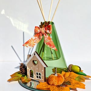 May include: A green glass cone-shaped reed diffuser with three wooden reeds. The bottle is decorated with an orange and yellow leaf-patterned ribbon, a small wooden house cutout, a pinecone, an acorn, and several artificial autumn leaves.