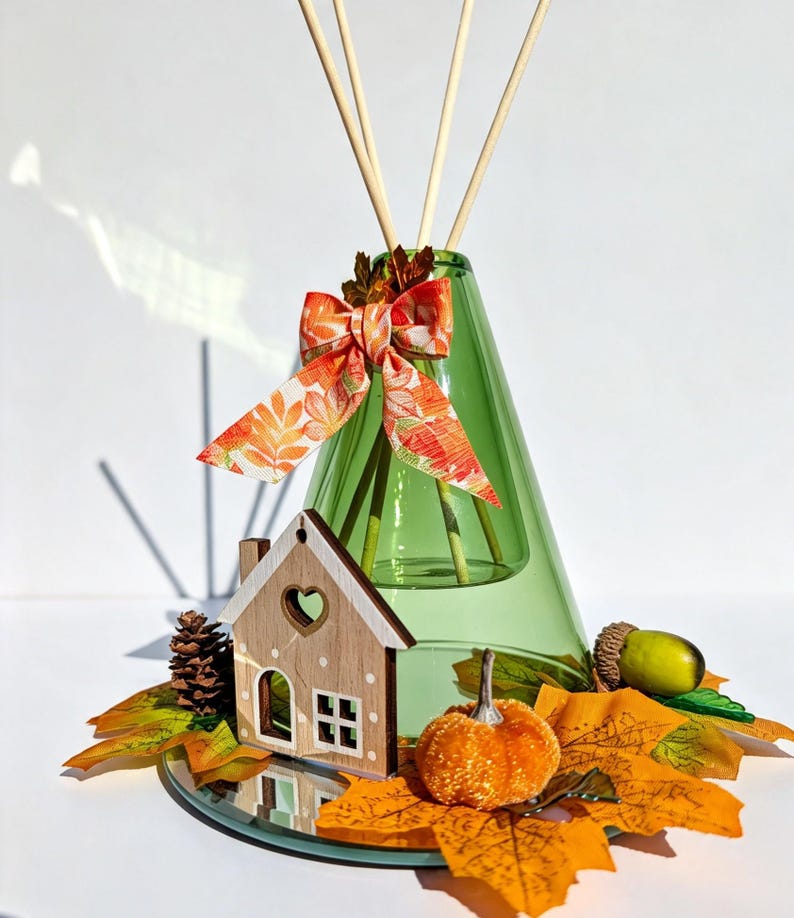 May include: A green glass cone-shaped reed diffuser with three wooden reeds. The bottle is decorated with an orange and yellow leaf-patterned ribbon, a small wooden house cutout, a pinecone, an acorn, and several artificial autumn leaves.