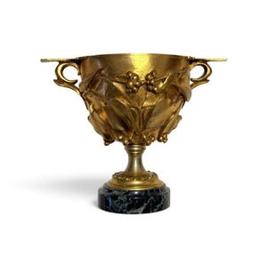Könnte beinhalten: Ein goldfarbener Metallpokal mit kunstvollen Reliefs aus Blättern und Beeren an der Außenseite der Schale. Das Gefäß verfügt über zwei seitliche Griffe und wird von einem Sockel getragen, der auf einem runden, dunkelgrünen Marmorfuß steht.