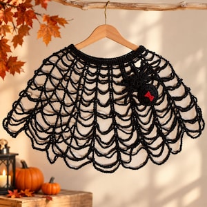 May include: A black crocheted capelet featuring an intricate spiderweb pattern. The garment is constructed with a circular neckline and a scalloped hem. A three-dimensional black crocheted spider with a small red bow detail is attached to the web.