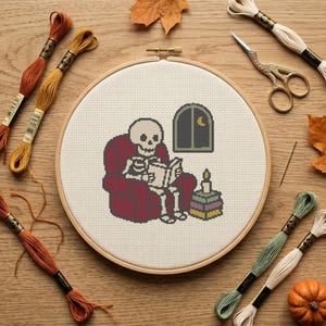 Puede incluir: Un bastidor de bordado circular de madera con un patrón de punto de cruz de un esqueleto sentado en un sillón rojo leyendo un libro. Junto al esqueleto hay una pila de libros con una vela encendida y una ventana con una luna creciente.